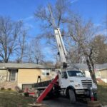 A PrimeCut Tree Service crane truck parked in an Omaha driveway, extending its boom over a house for a large-scale winter tree removal.