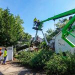PrimeCut Tree Service crew in a green boom lift maneuvering a large log during a residential tree removal next to a house in Omaha.